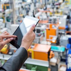Horizontal color image of businesswoman - unrecognizable person - working with digital tablet in large futuristic factory. Woman standing on top of a balcony, holding touchpad and checking inventory of a manufacturing company on touchscreen tablet. Focus on businesswoman's hands holding black tablet, futuristic machines in background.
